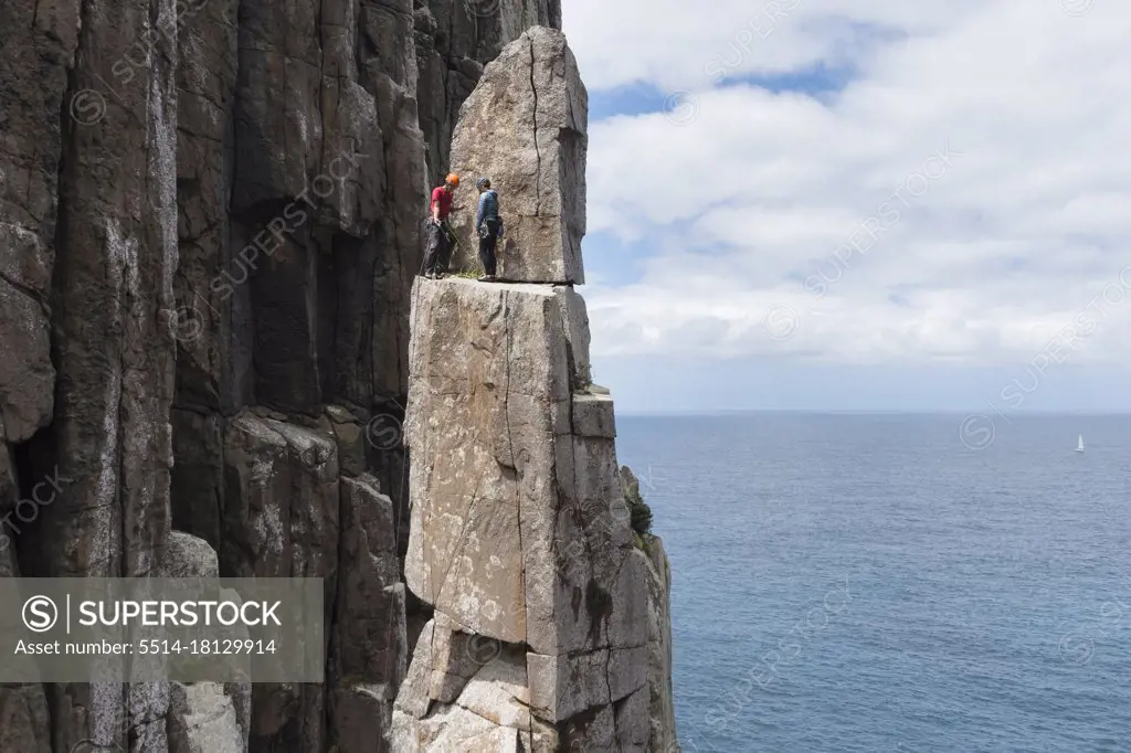 Adventurous couple summits the exposed and frail sea stack of the Totem Pole in a sunny day, with a sea cliff, the ocean and a sailing boat seen in the background in Cape Hauy, Tasmania