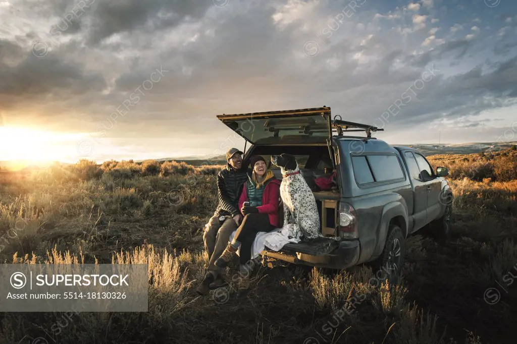 Cheerful couple sitting with dog at trunk of off-road vehicle on field during vacation