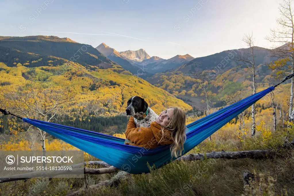 Happy young woman with dog relaxing on hammock in forest during vacation