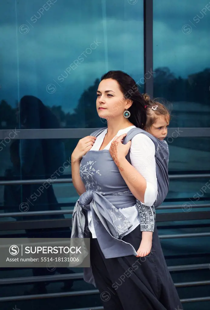 Mother and baby girl sit back in grey wrap sling stay near urban glass