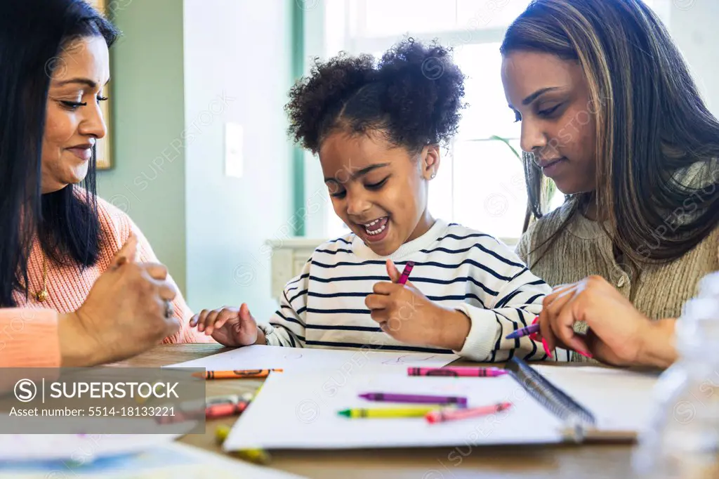 Mother and grandmother with cheerful girl drawing with crayon on paper at home