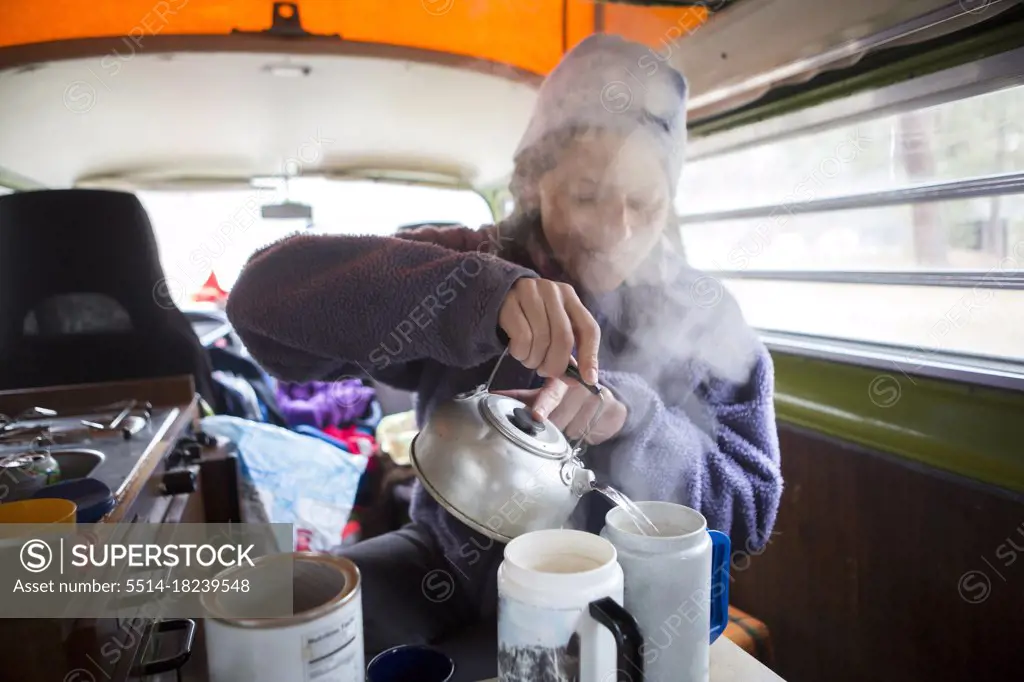 A woman pours boiling water for tea in VW camper van during road trip.