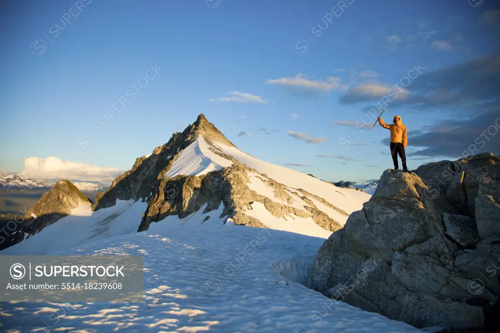 Successful climber wearing yellow jacket stands on rocky outcrop.