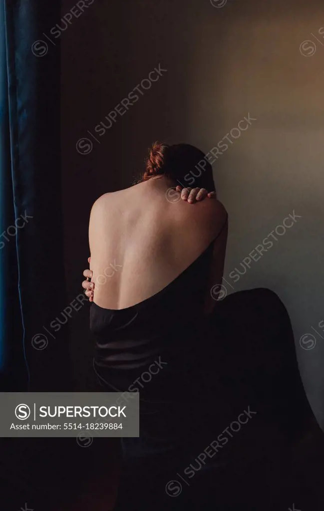 Back view of sad woman bent forward with back exposed in a dark room.