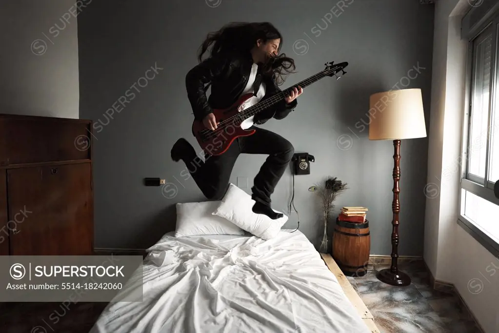 A man with long hair playing bass guitar on the bed