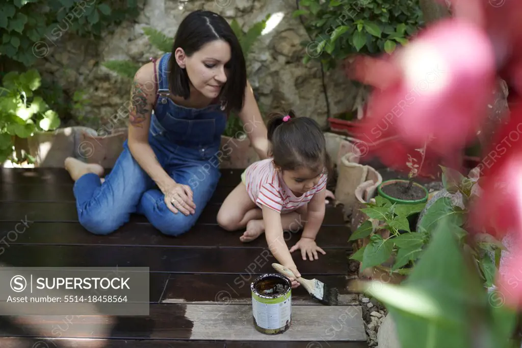 A mother watches as her little girl paints the wood in the garden.