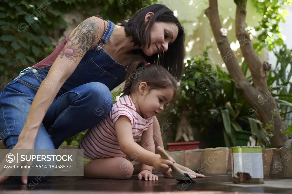 A girl enjoys painting the wood on the floor with her mother.