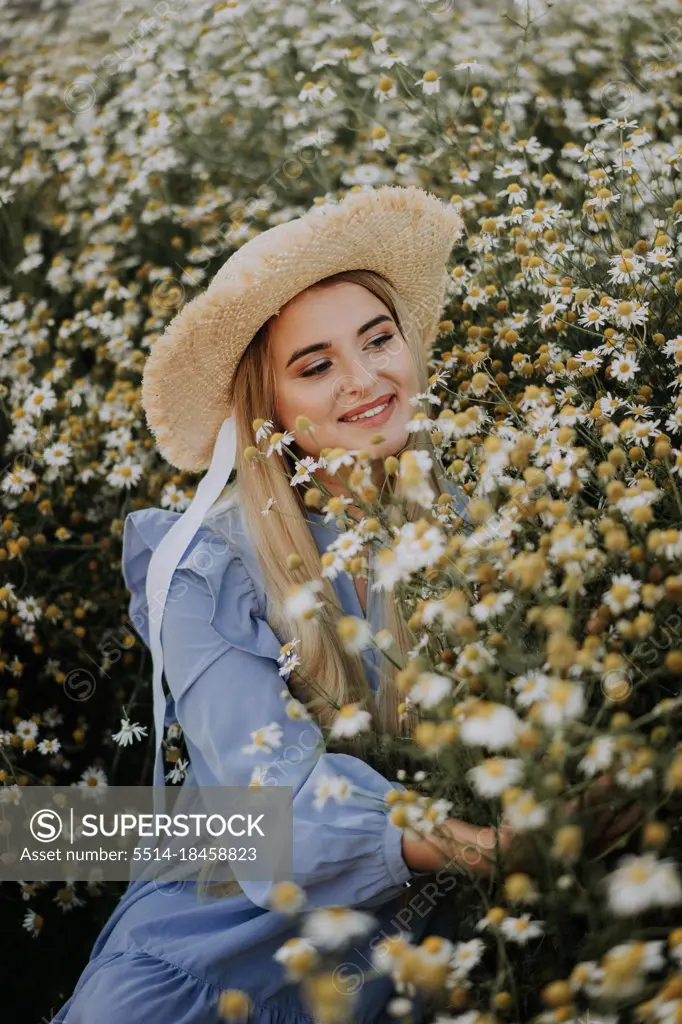 A young woman in straw hat is sitting in a chamomile field.