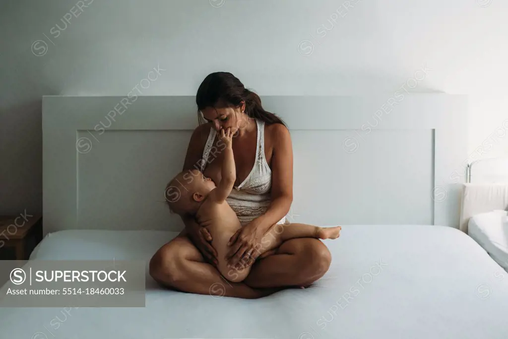 Mom breastfeeding while naked baby touches her face in bedroom