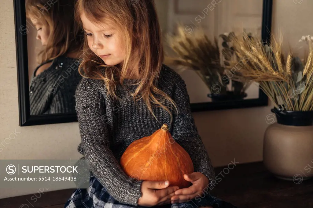 The cute girl holding a pumpkin in her hands sitting on the table.