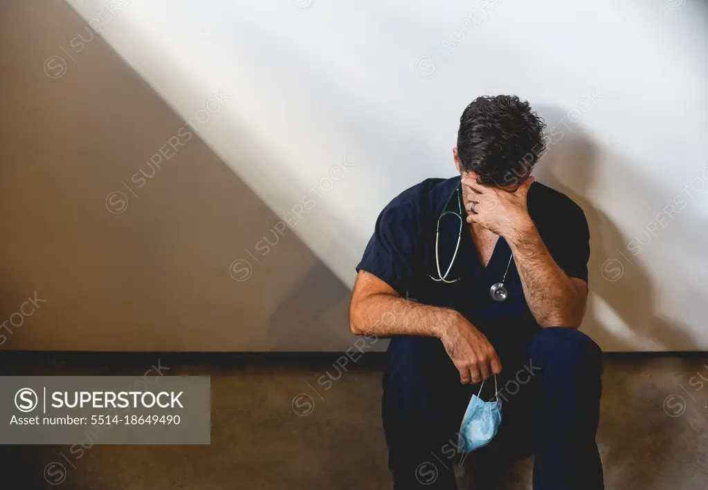 Exhausted doctor wearing scrubs sitting on floor in a patch of light.