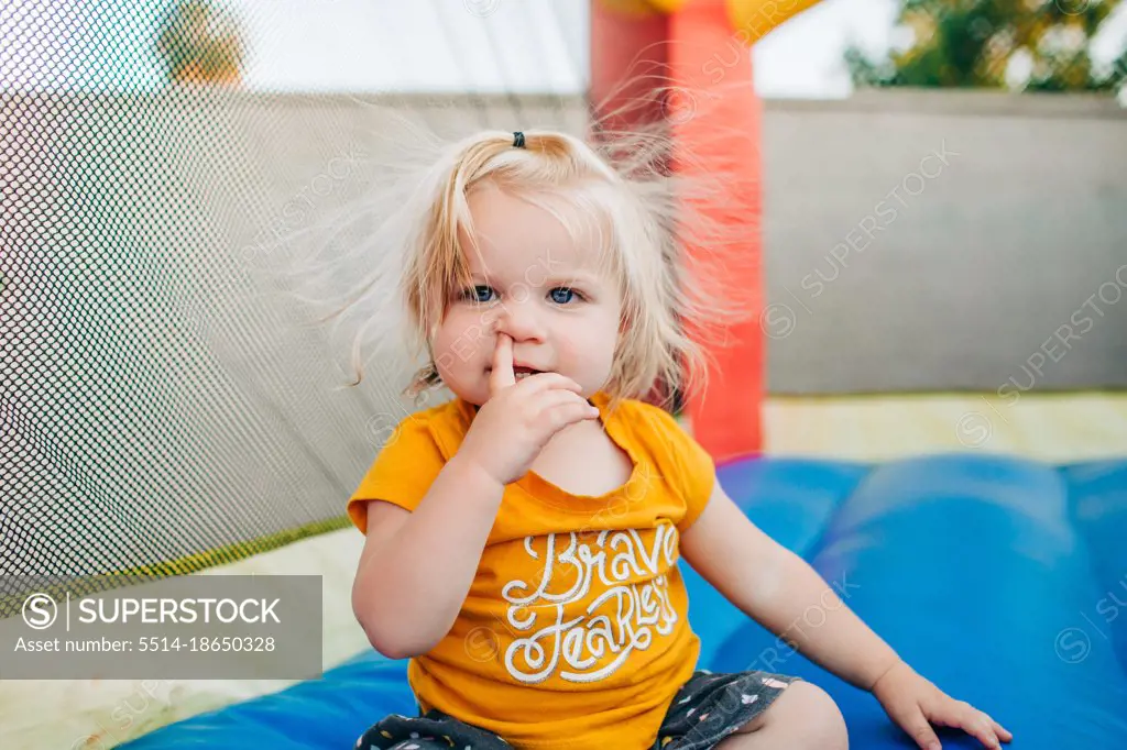 Blonde toddler girl picks nose while playing on jump house in backyard
