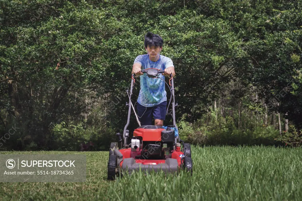 Asian teen boy in tie dyed shirt cuts grass pushing red lawn mower