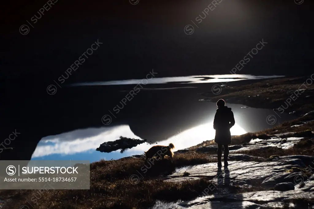Silhouette of woman and dog against mountain reflection in lake