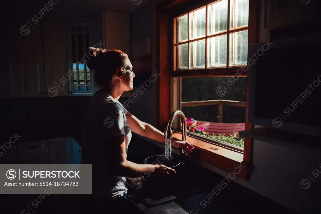 Woman filling a pot at the sink under a window in a dark kitchen.