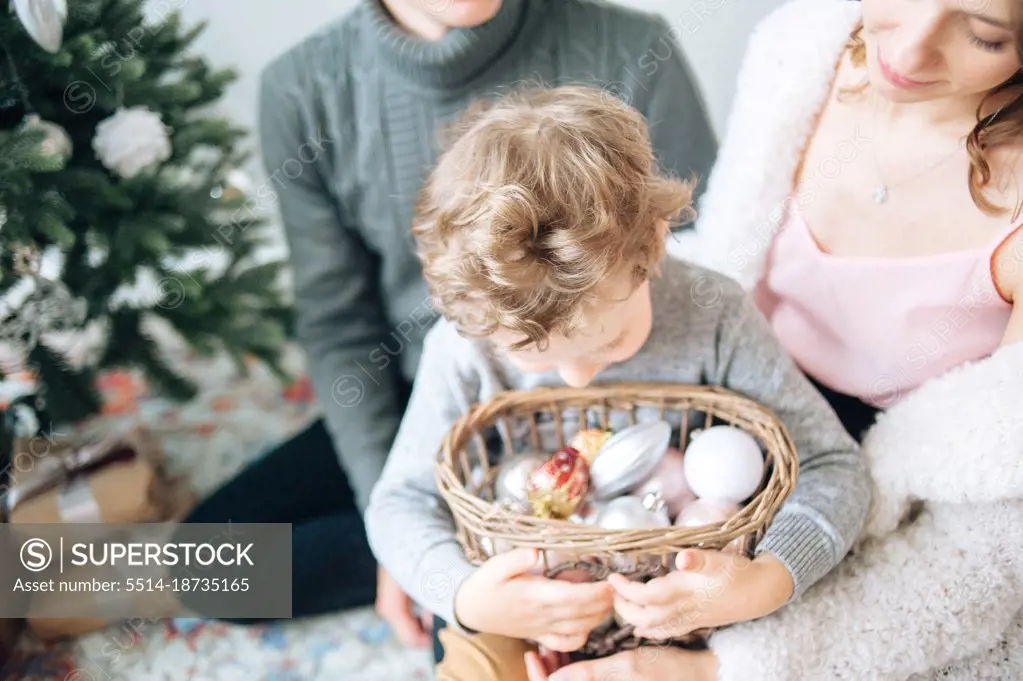 Curly boy hugs basket with Xmas decor while sitting on mother legs