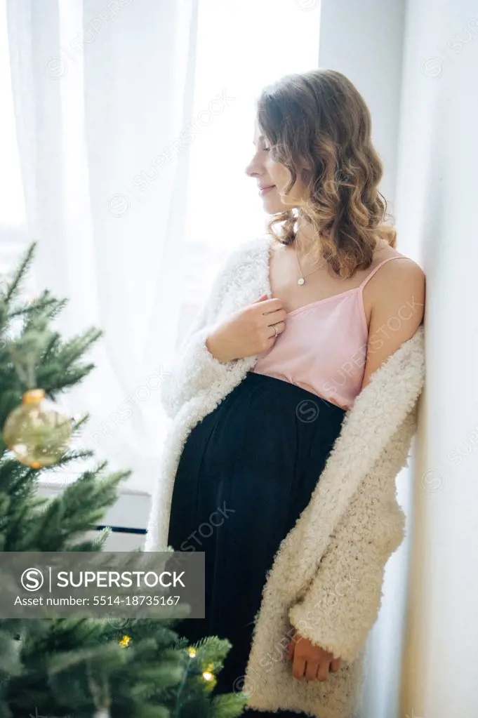 Beautiful pregnant woman stands by the window beside Christmas tree