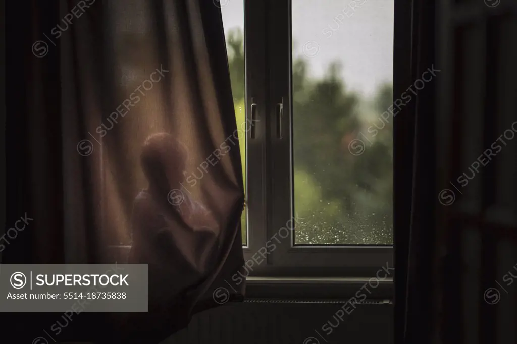 Child sitting on windowsill and hiding behind brown curtain