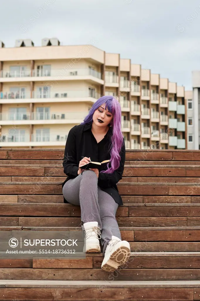 Non-binary person with purple hair writes in a notebook on the street