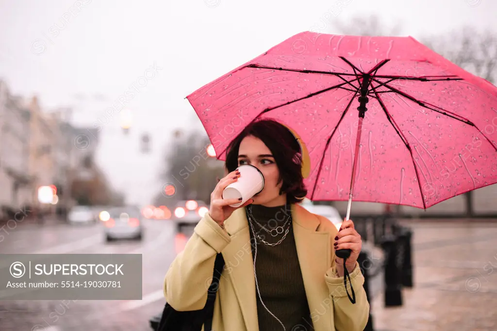 Young woman with an umbrella drinking coffee walking in city