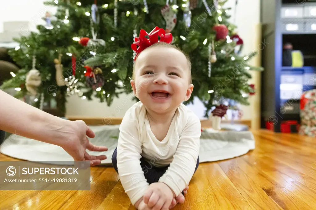 Happy Baby Sits on Floor Next to Christmas Tree with Bow on Head