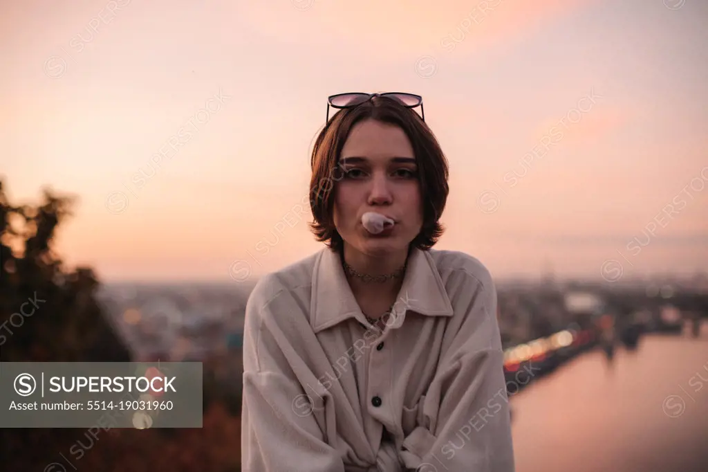Young woman blowing bubble gum against city at sunset