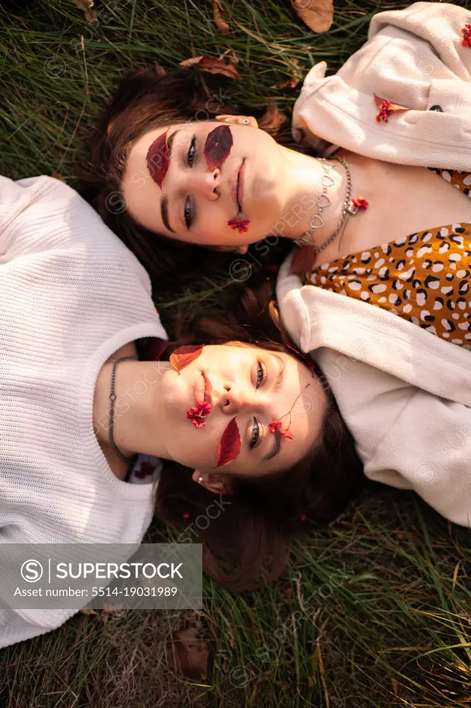 Two teenage girls with red leaves on faces lying on grass in autumn