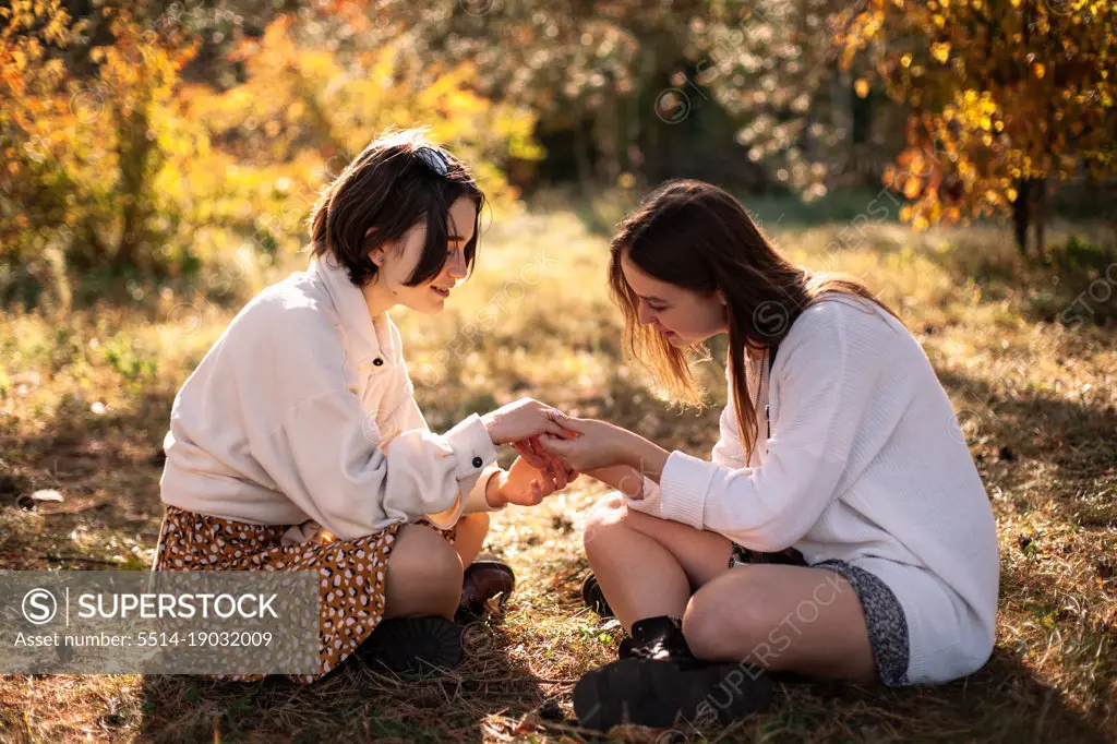 Girlfriends holding hands while sitting on grass in park in autumn