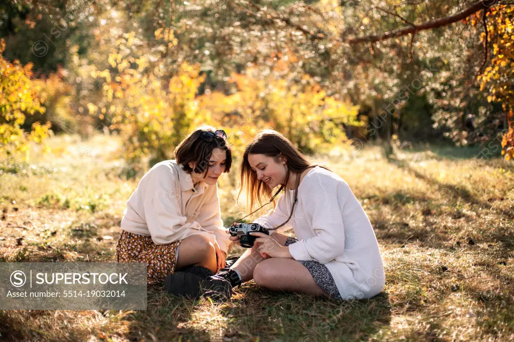 Two teenage girls with camera sitting on grass in park in autumn