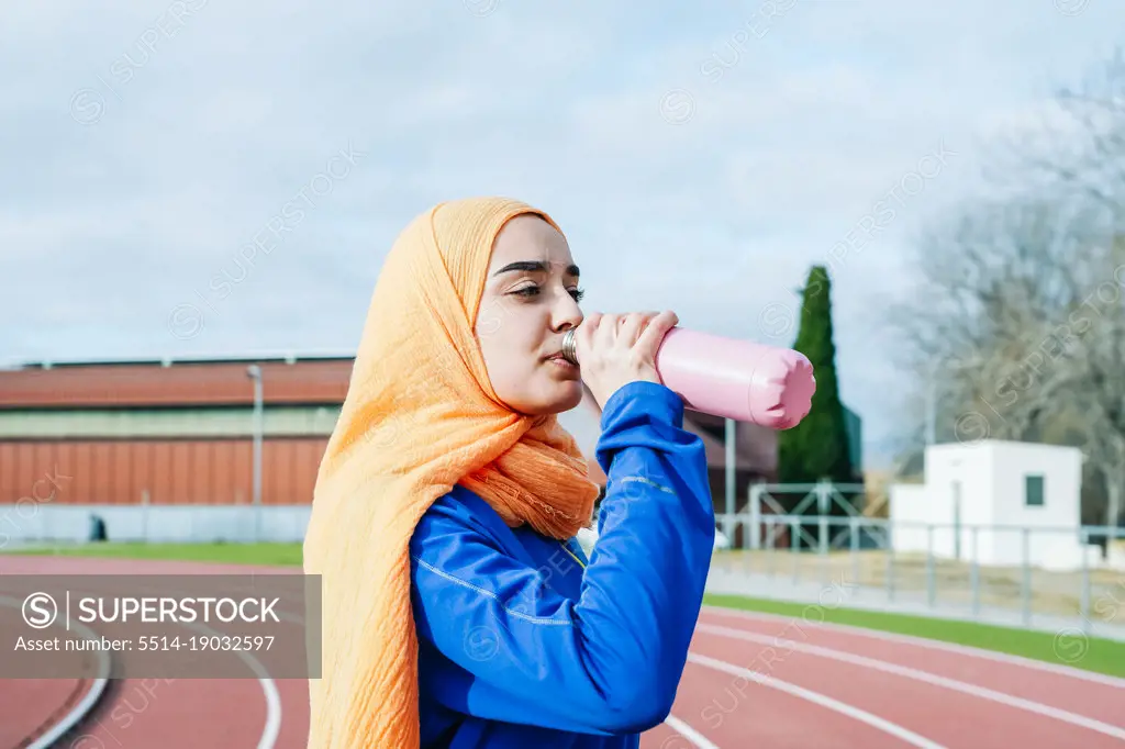 Ethnic woman drinking water after running training on racetrack