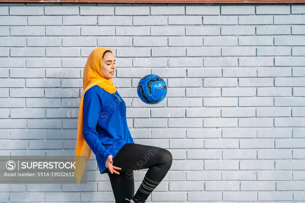 Ethnic woman kicking ball with knee near brick wall