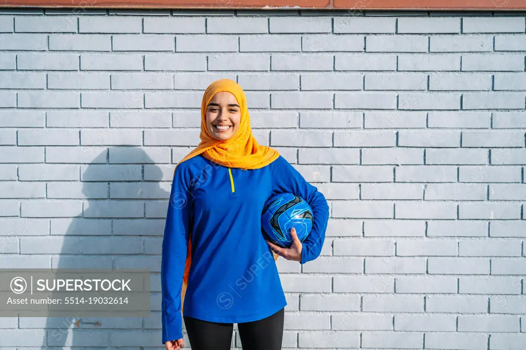 Cheerful Islamic woman with ball near brick wall
