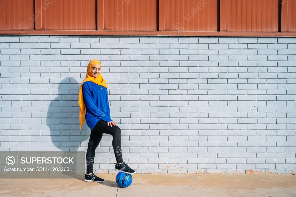 Smiling ethnic football player with leg on ball near brick wall