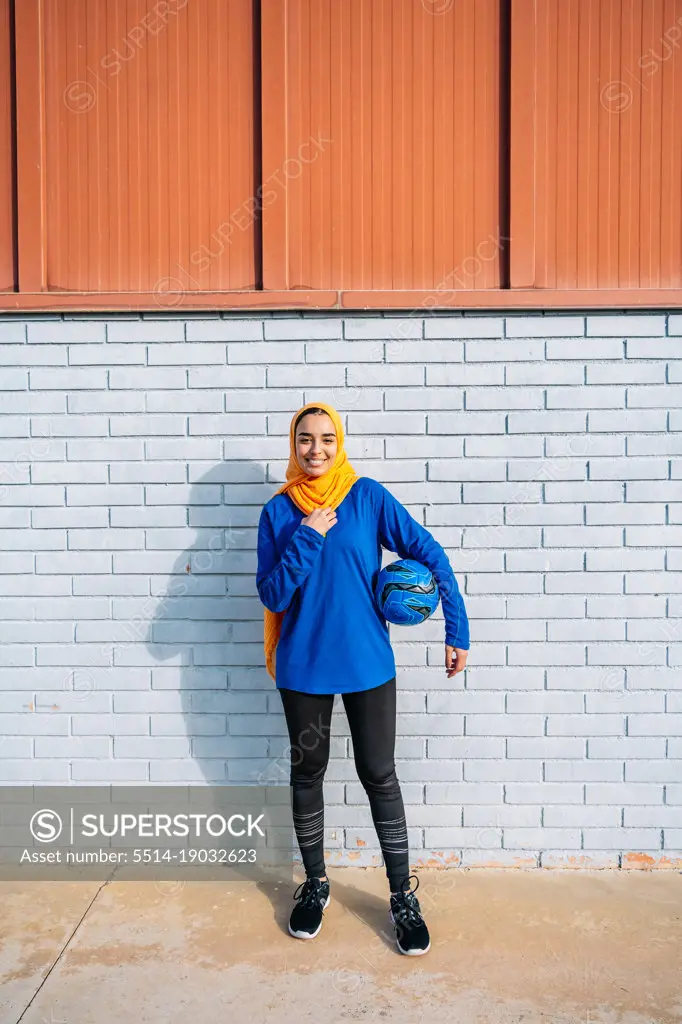 Smiling ethnic woman with ball for training near brick wall