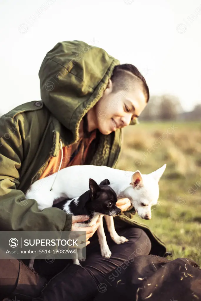 androgynous person sits in sunny field with two chihuahuas smiling