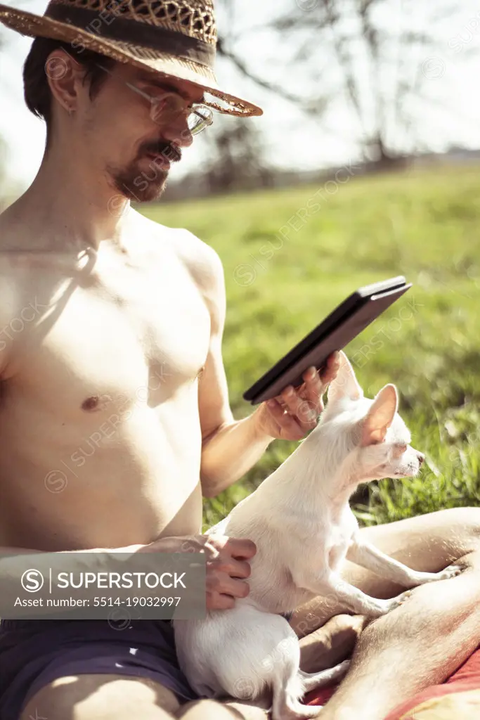 Sunbaking man reads kindle with dog on lap on grass meadow