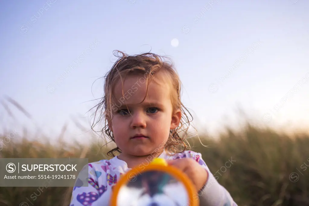 Young child holds flashlight and looks at camera with grasses behind