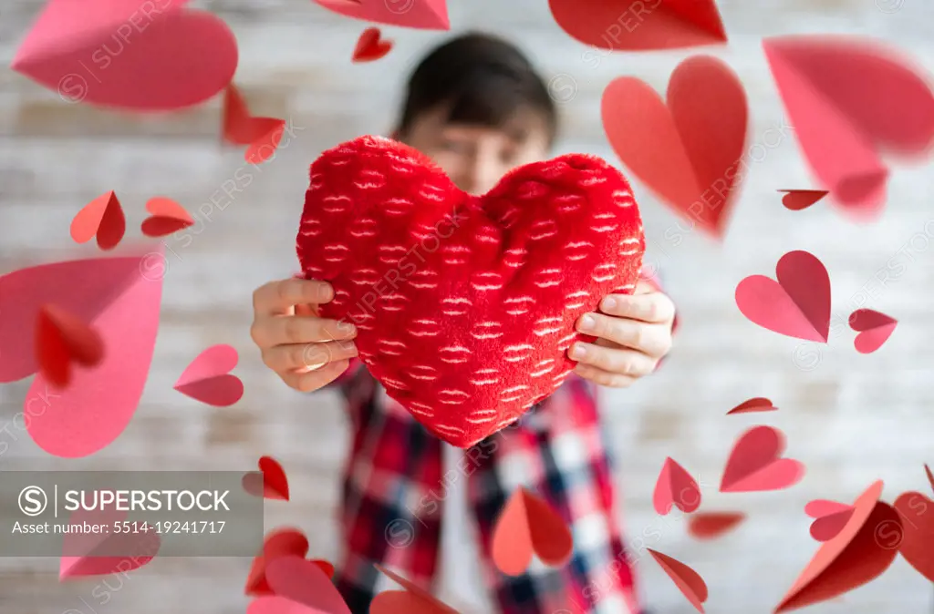 Boy holding heart pillow surrounded by floating paper hearts.