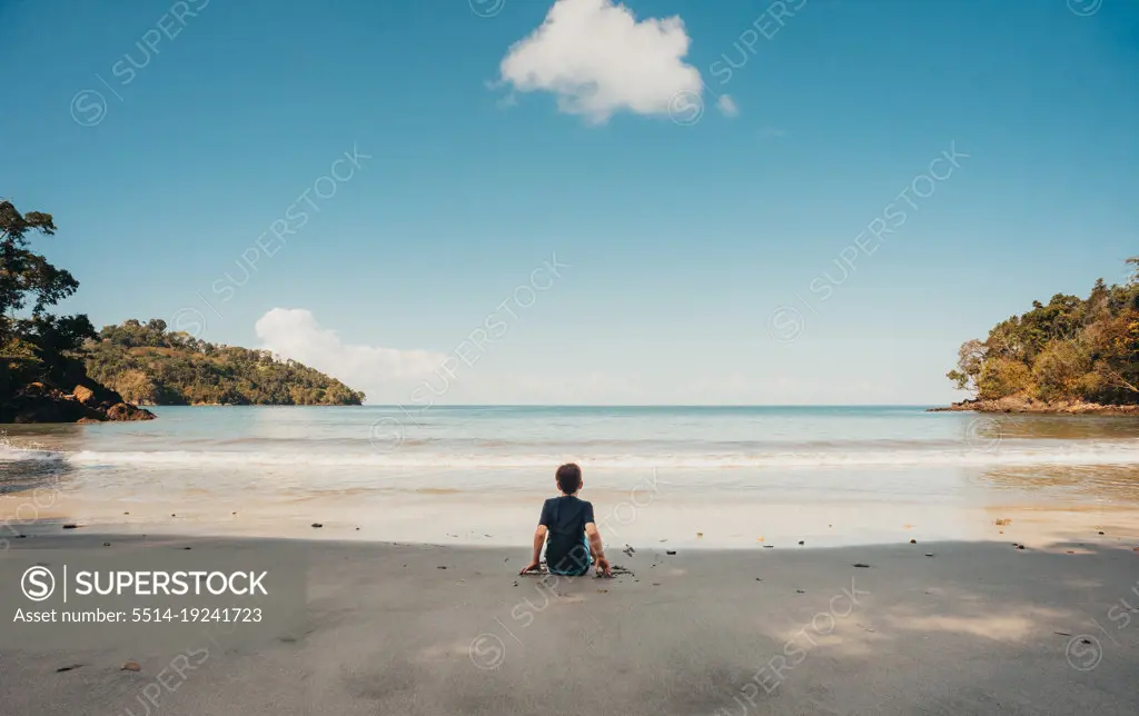 Boy sitting alone on tropical beach looking at the ocean on sunny day.