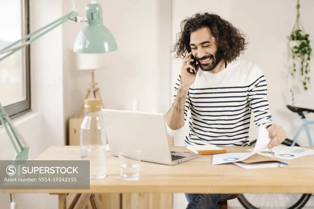 Cheerful young man talking on phone while working from home