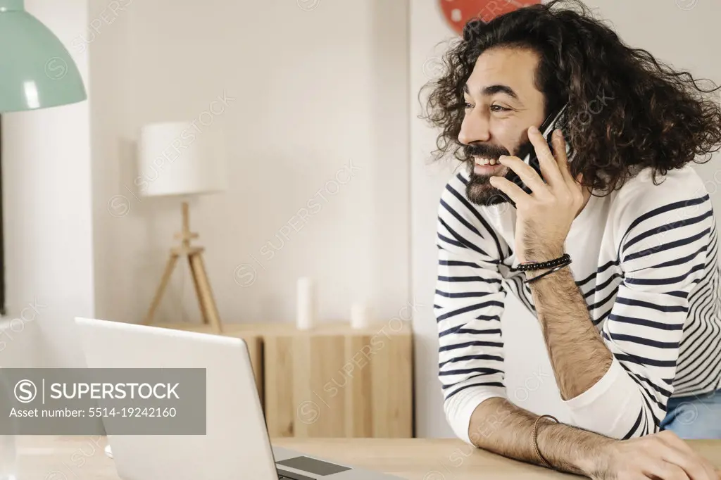 Cheerful man talking on phone with his laptop next to him at home
