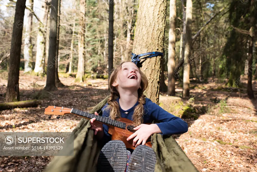 child sat in a hammock playing the ukulele in the forest