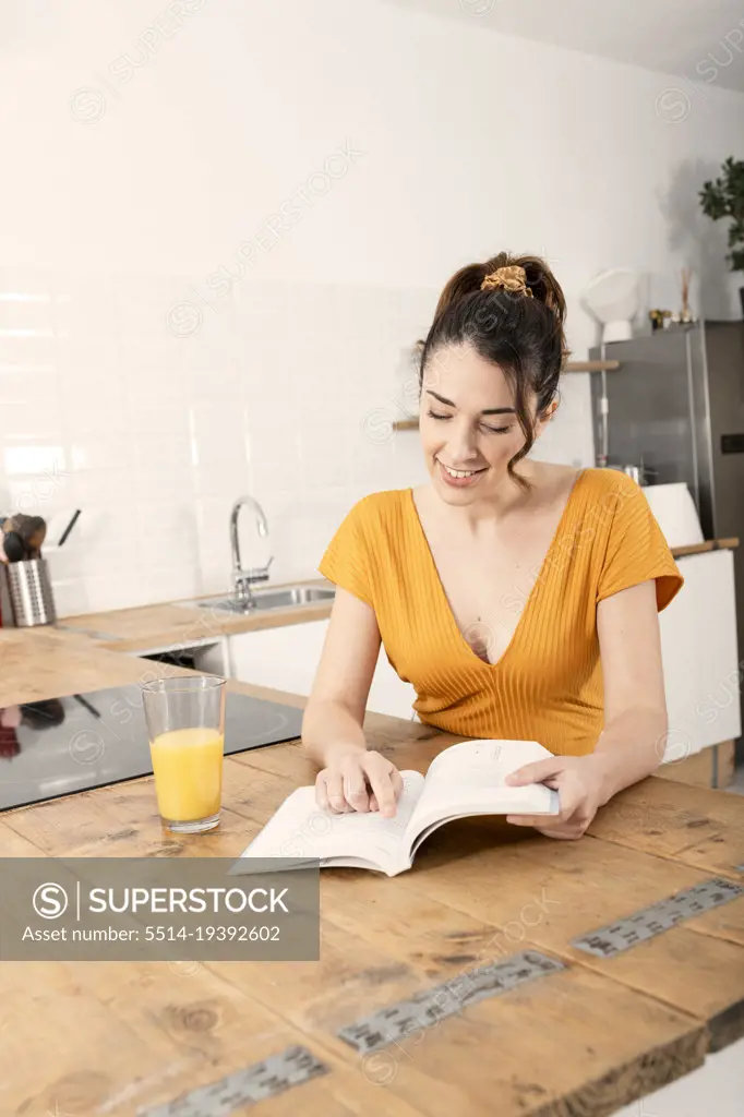 Beautiful young woman reading book in kitchen near orange juice