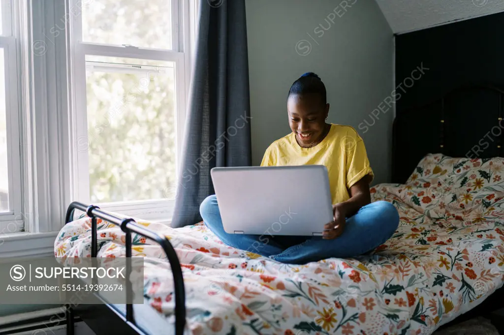 Young black girl using laptop while sitting on bed in her room