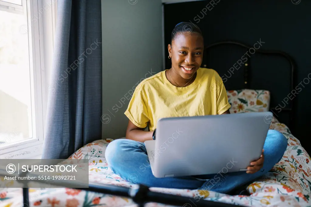 Happy black teenage girl sitting on bed with her computer