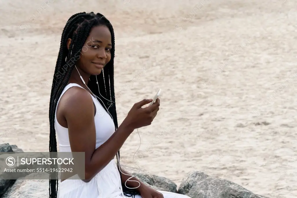Woman with braids with headphones sitting at the beach using pho
