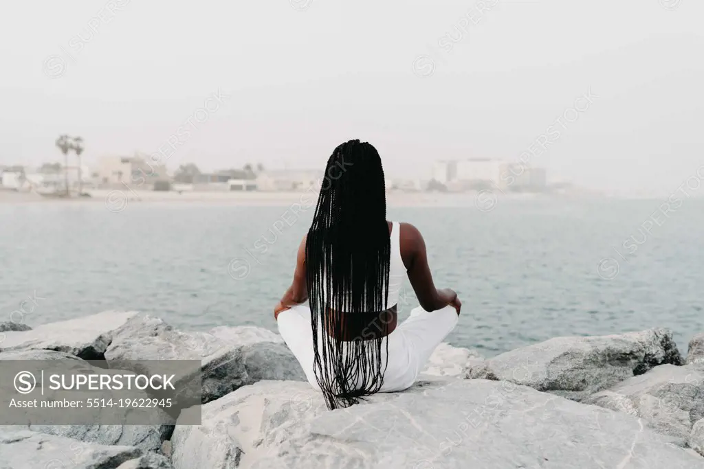 Woman with braids sittingon the rocks at the beach meditating