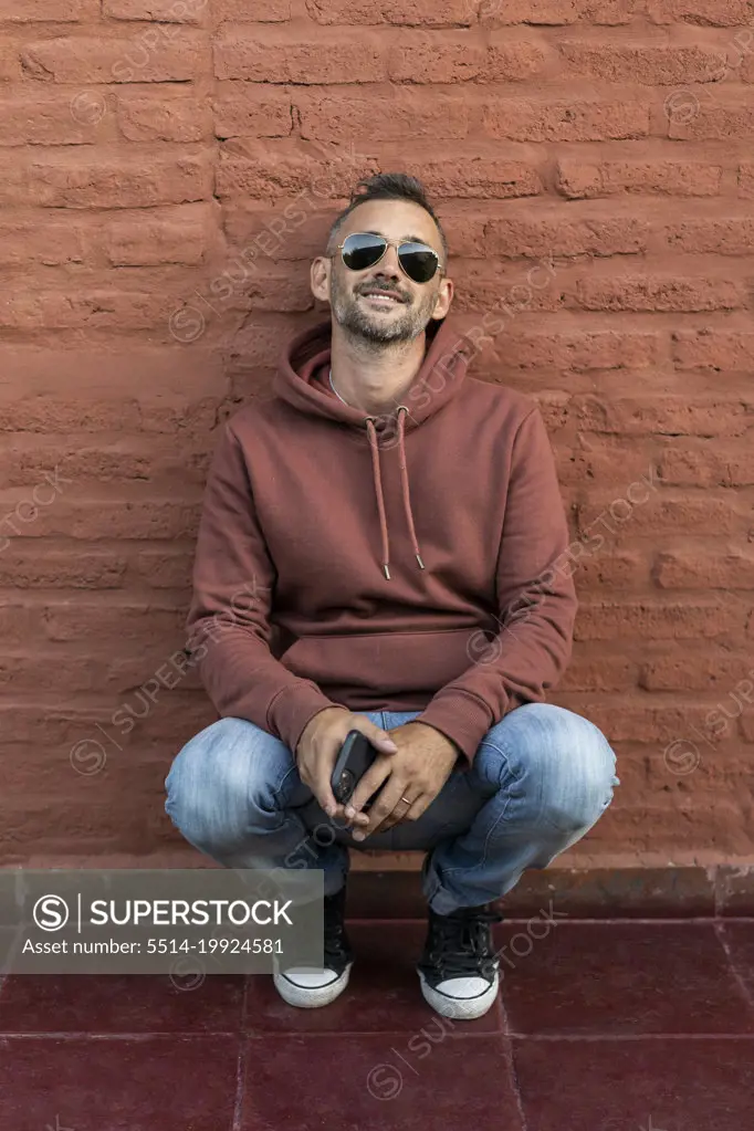 A handsome man crouching while leaning on a red brick wall
