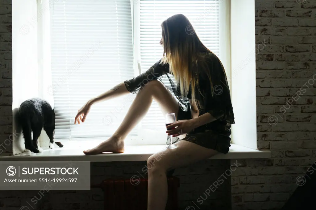 Woman in nightgown sitting on windowsill with glass of wine