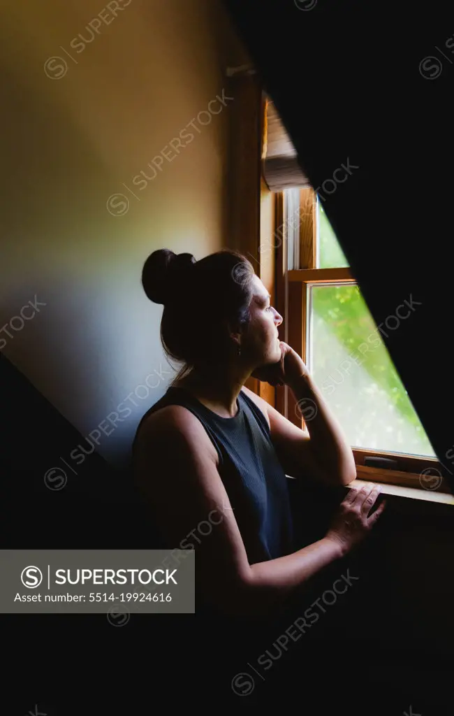 Woman looking out of a window in a dark room in the daytime.